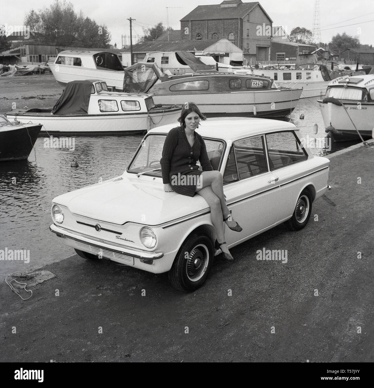 1967, historical, outside at a boatyard, we see a young lady wearing a woollen cardigan and skirt sitting or posing in the boot of a Sunbeam Sport motorcar,  the sports version of the famous small car, the HIllman Imp. The engine was at the rear and so the boot was at the front. The Hillman Imp was a rear-engined small car made by Rootes Group and its successor Chrysler Europe from 1963 until 1976. It was the competitor in the small car category to the British Leylands Mini. Stock Photo