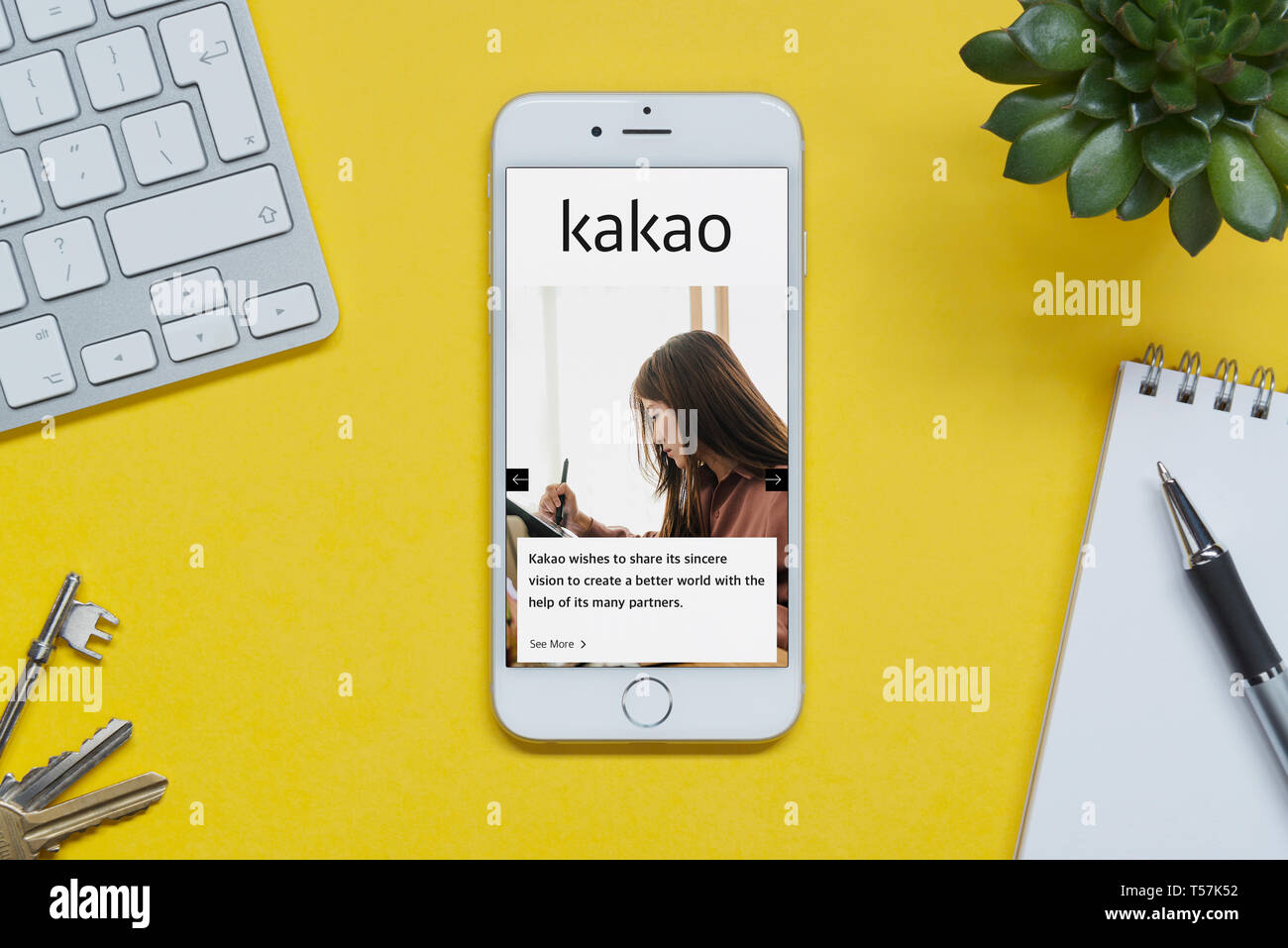 An iPhone showing the Kakao website rests on a yellow background table with a keyboard, keys, notepad and plant (Editorial use only). Stock Photo