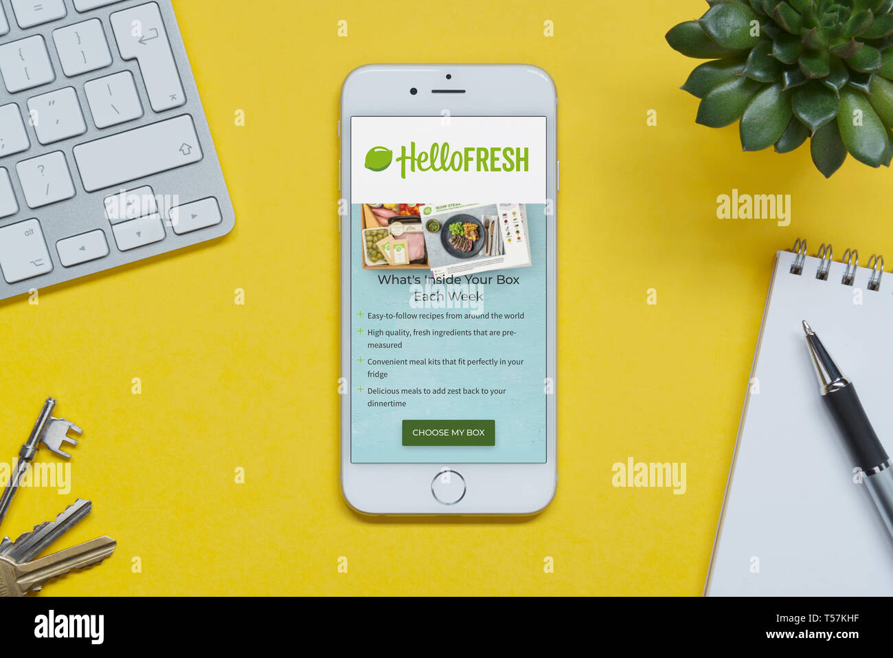 An iPhone showing the Hello Fresh website rests on a yellow background table with a keyboard, keys, notepad and plant (Editorial use only). Stock Photo