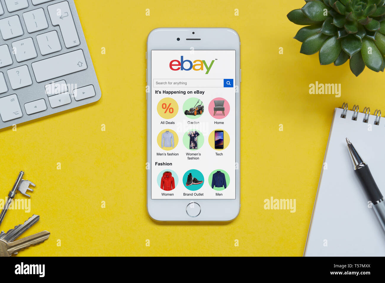 An iPhone showing the ebay website rests on a yellow background table with a keyboard, keys, notepad and plant (Editorial use only). Stock Photo