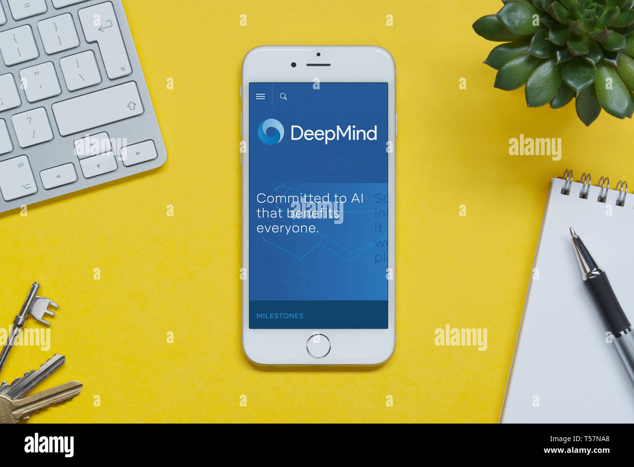 An iPhone showing the DeepMind website rests on a yellow background table with a keyboard, keys, notepad and plant (Editorial use only). Stock Photo