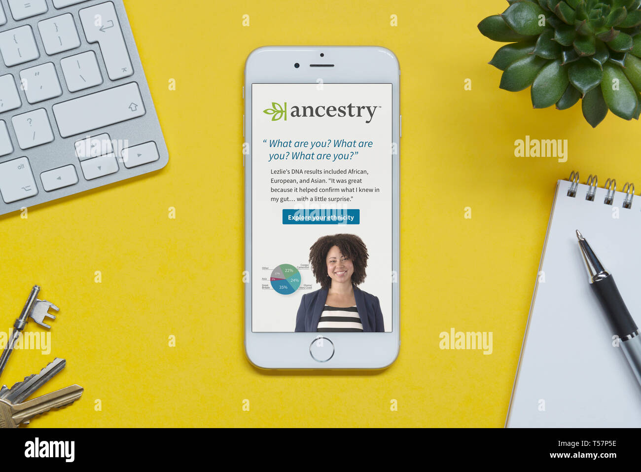 An iPhone showing the Ancestry website rests on a yellow background table with a keyboard, keys, notepad and plant (Editorial use only). Stock Photo