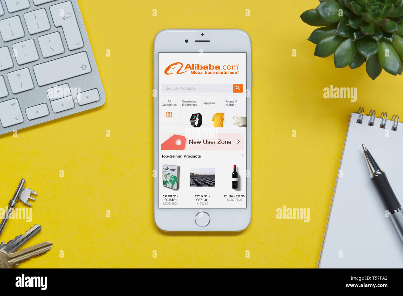 An iPhone showing the Alibaba website rests on a yellow background table with a keyboard, keys, notepad and plant (Editorial use only). Stock Photo