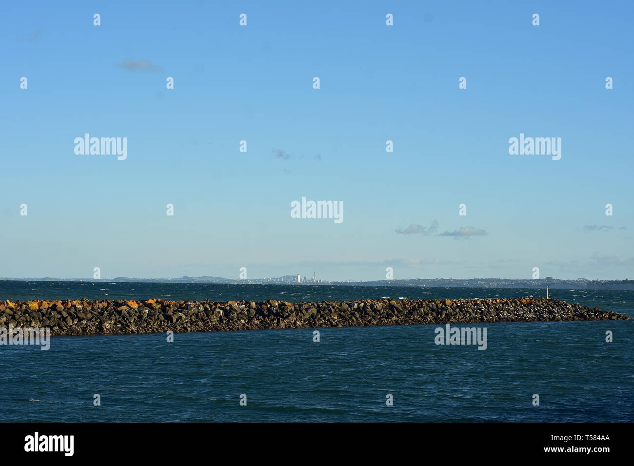 Breakwall of free laid stones surrounded by dark choppy sea with Auckland cityscape in distant background. Stock Photo