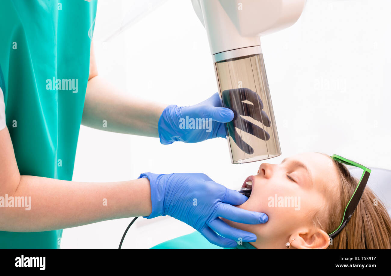 Radiographer taking teeth radiography to elementary age girl using digital x-ray machine in pediatric dental clinic. Doctor pointing X-ray unit while  Stock Photo