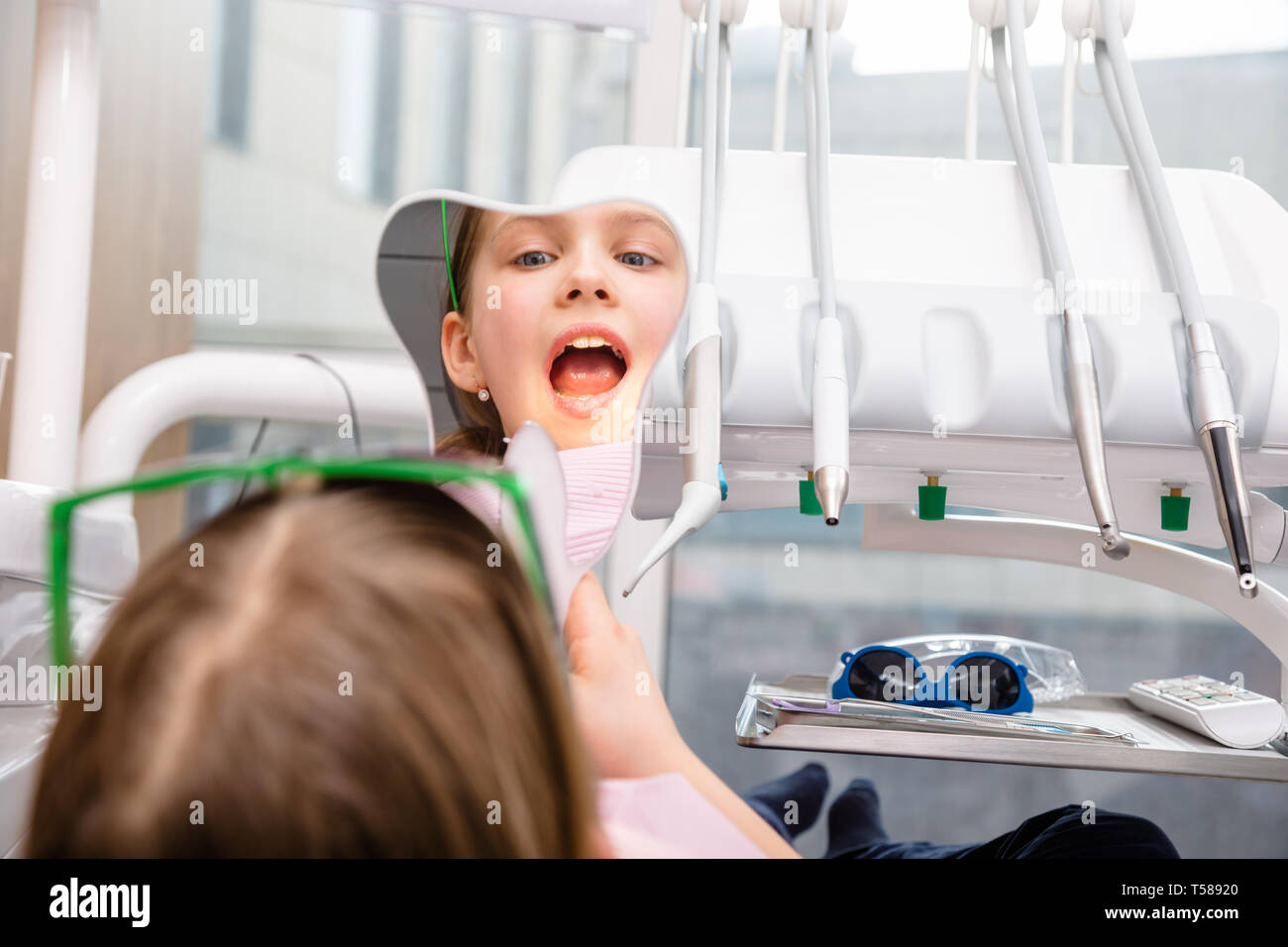 Preteen girl sitting in a dental chair inspecting repaired teeth looking at tooth-shaped mirror in pediatric dental clinic Stock Photo