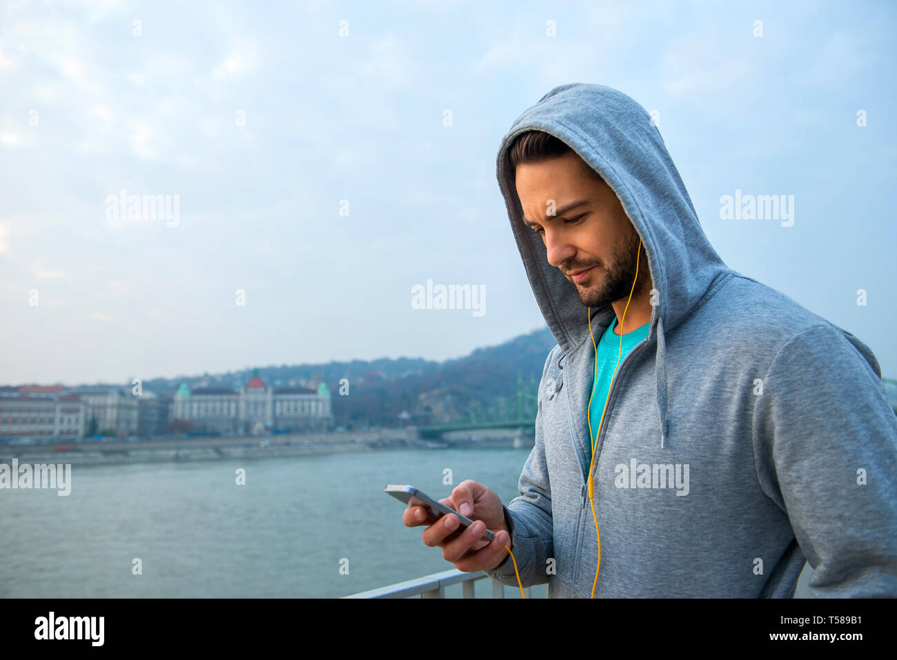 A handsome young man standing in training clothes and using his phone with earphones at the riverside Stock Photo