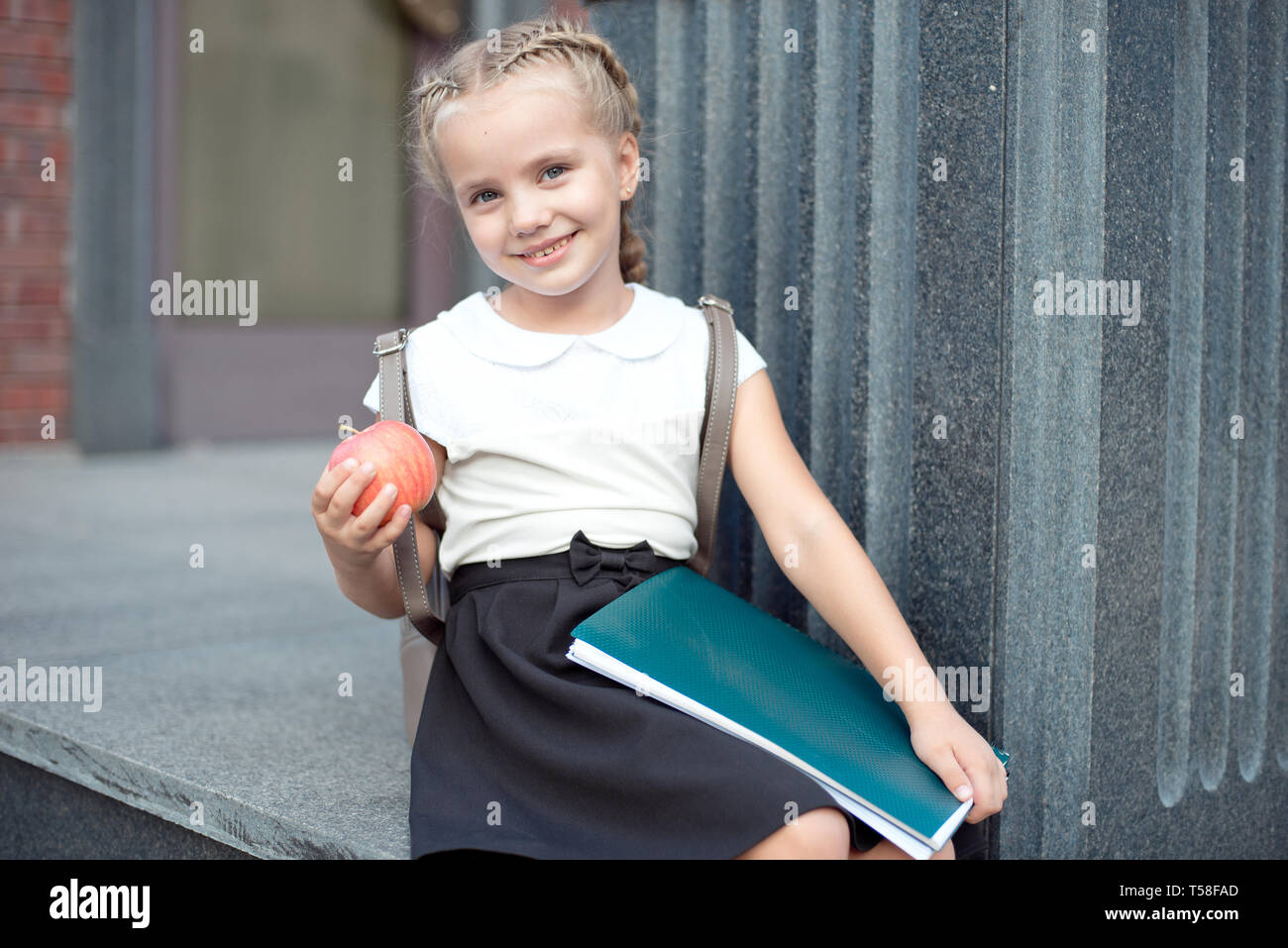 Happy little schoolgirl with blond hair in school uniform with lunch, book sits near school Stock Photo