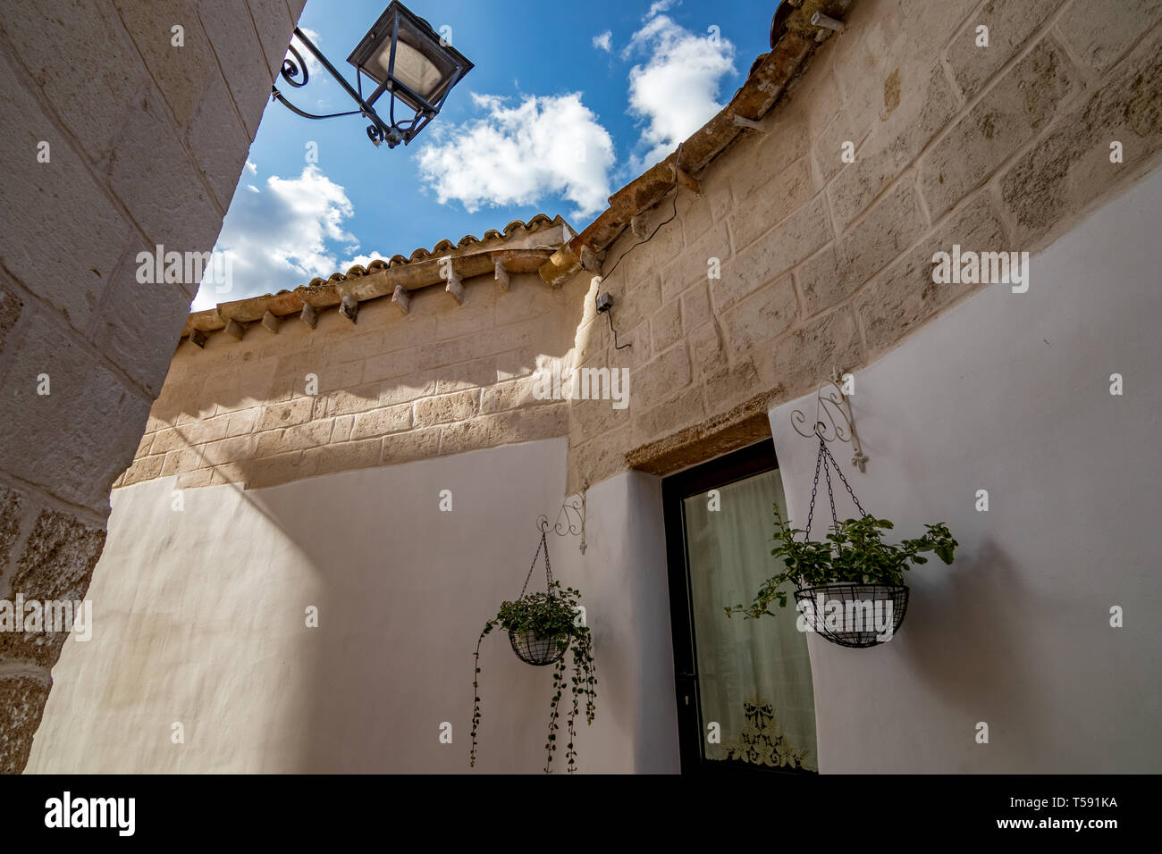 Two green potted plants grow hanging in two white pots around glass door in Laterza, Puglia region, Southern Italy in summertime, white wall background and puffy white clouds on blue sky Stock Photo