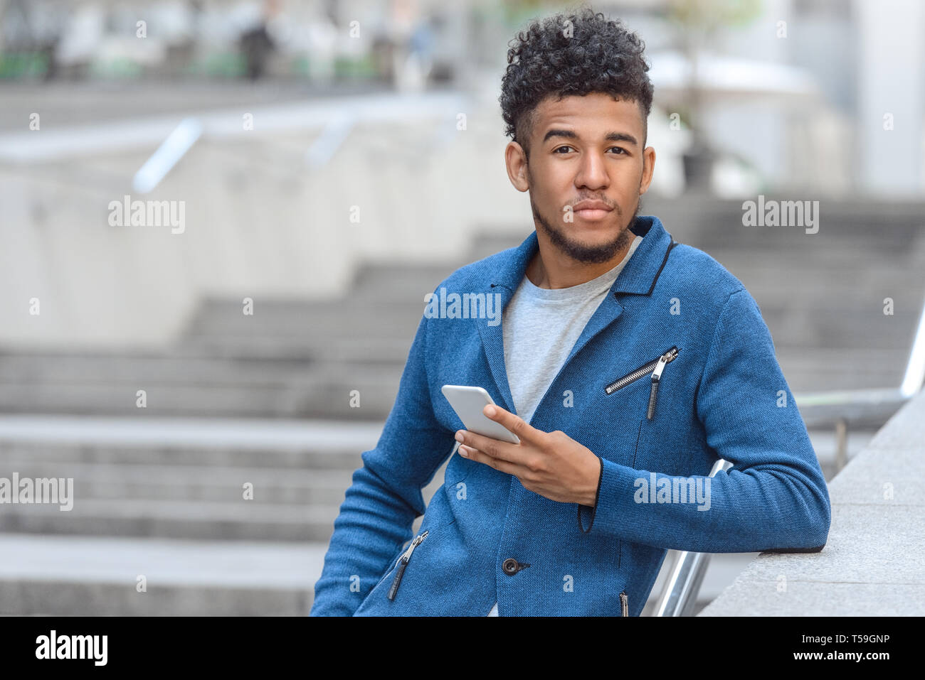 Young mulatto man standing leaning on concrete fence on city street holding smartphone looking camera concerned close-up blurred background Stock Photo