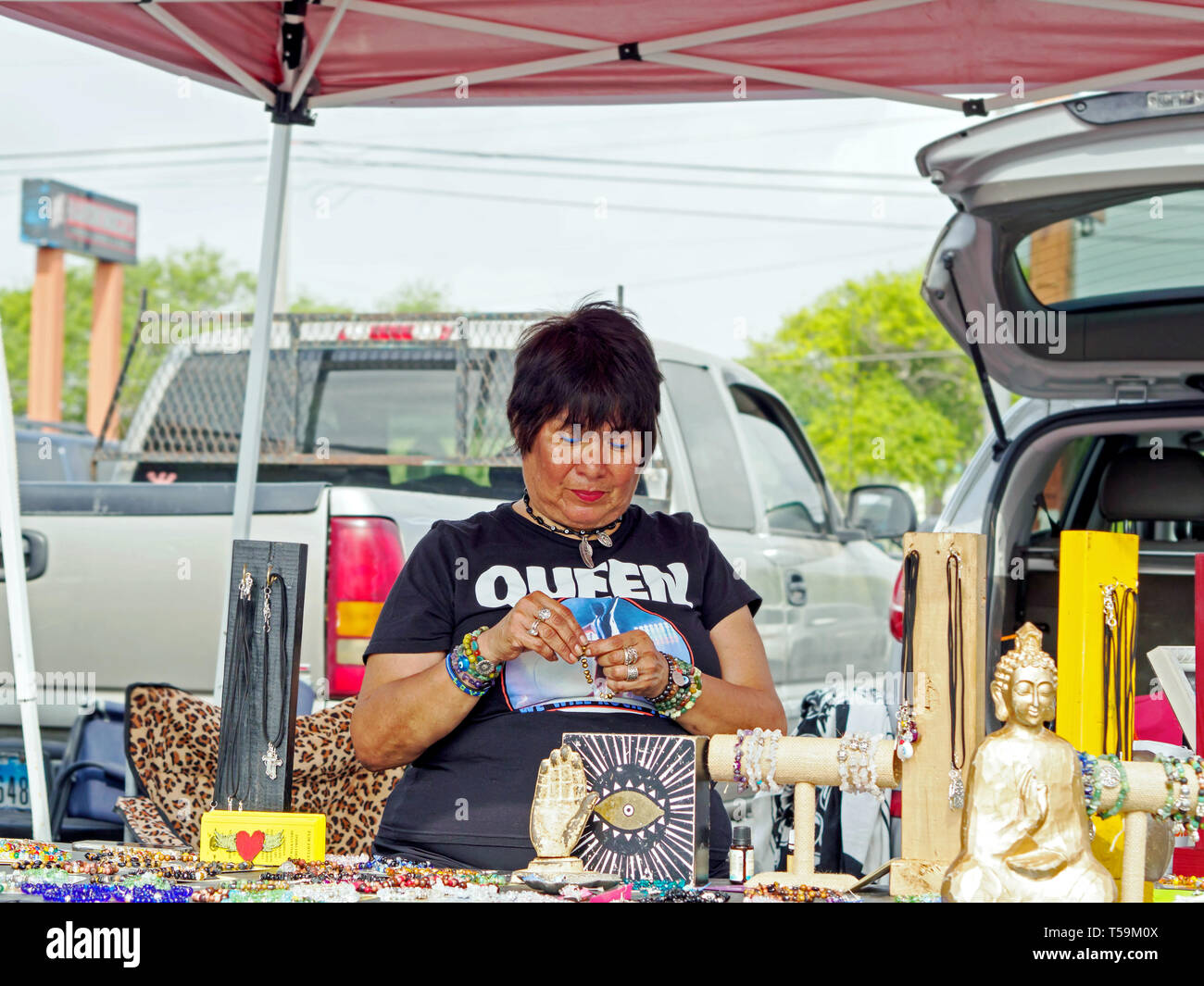 A dark-haired, female jewelry vendor wearing a 'Queen,' t-shirt, bracelets, and necklace staffs her booth. Corpus Christi Southside Farmers' Market. Stock Photo