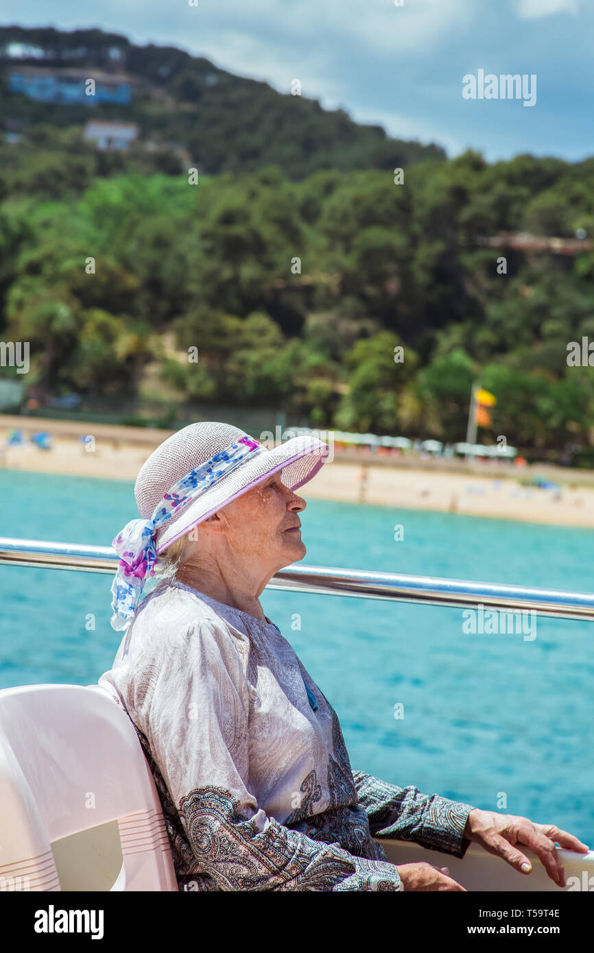 Elderly pretty woman in sun-hat enjoys summer vacation in a sea voyage on a pleasure boat. She sits on the upper deck Stock Photo
