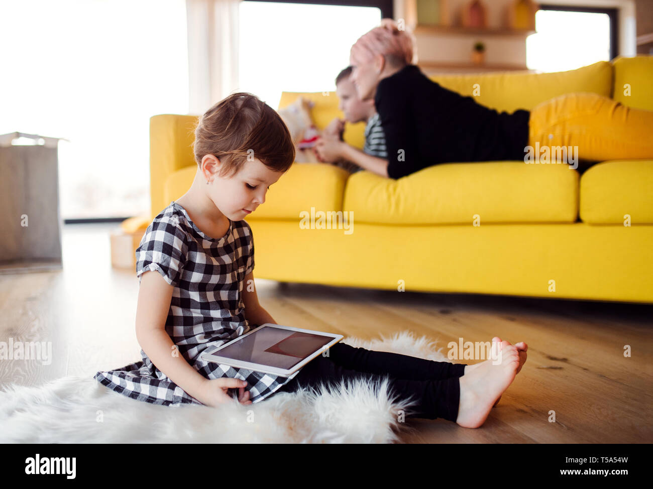 A small girl with mother and brother using tablet indoors on the floor at home. Stock Photo