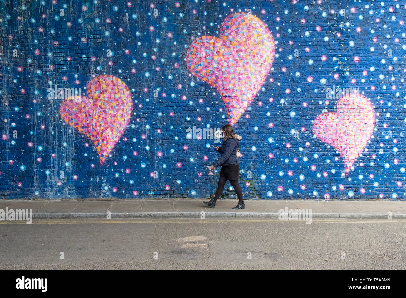 A woman looking at her mobile phone walking past a large painted mural of pink hearts in a street in London. Stock Photo