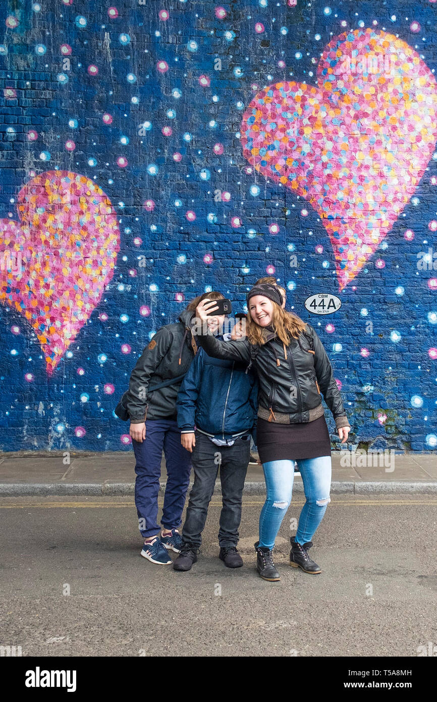 Tourists having fun taking a selfie in front of a large painted mural on a wall in a street in London. Stock Photo