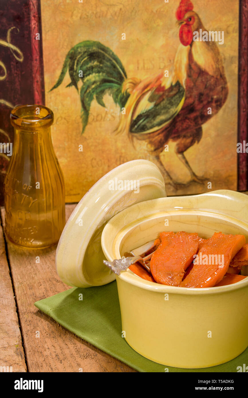 Baked candied sweet potatoes in an antique round casserole bowl resting on a cloth napkin, on a rustic wood tabletop with chicken artwork and an antiq Stock Photo