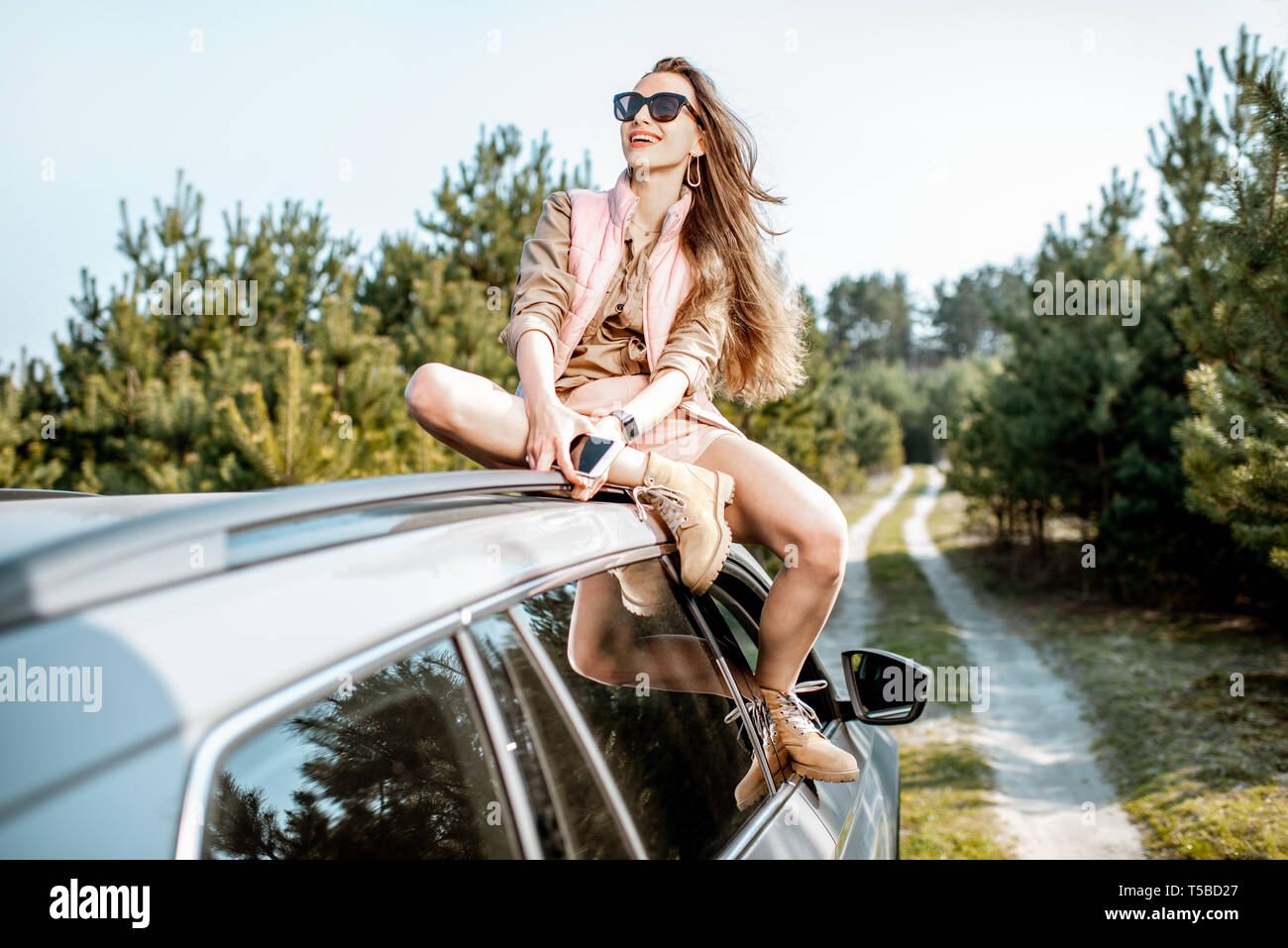 Young woman enjoying the trip while sitting on the car roof on a picturesque road in the woods Stock Photo