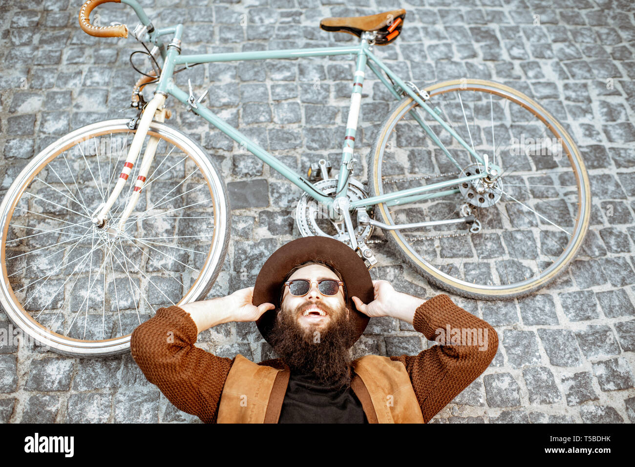 Portrait of a stylish bearded man in hat lying with retro bicycle on the pavement street, view from above Stock Photo