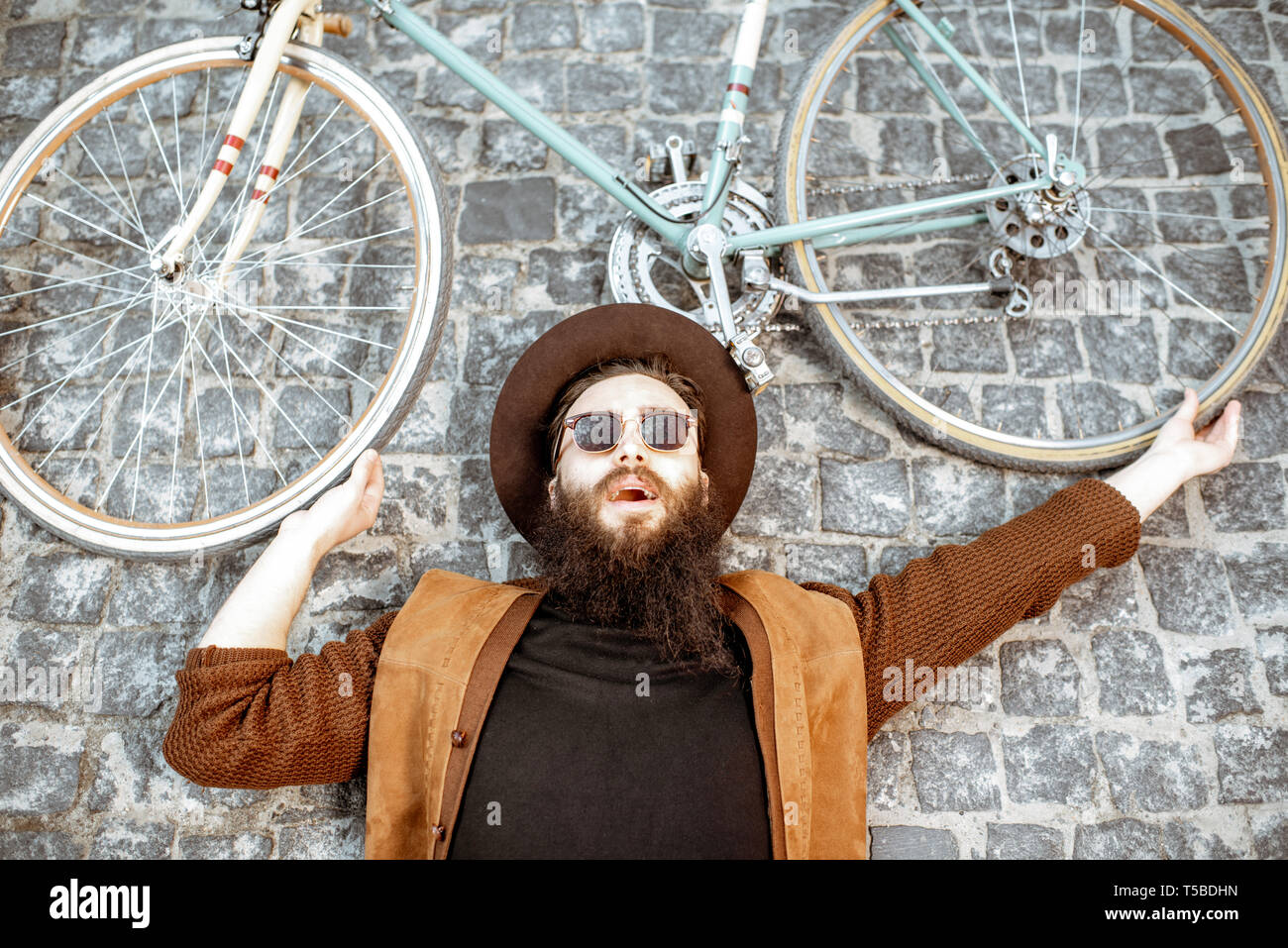 Portrait of a stylish bearded man in hat lying with retro bicycle on the pavement street, view from above Stock Photo