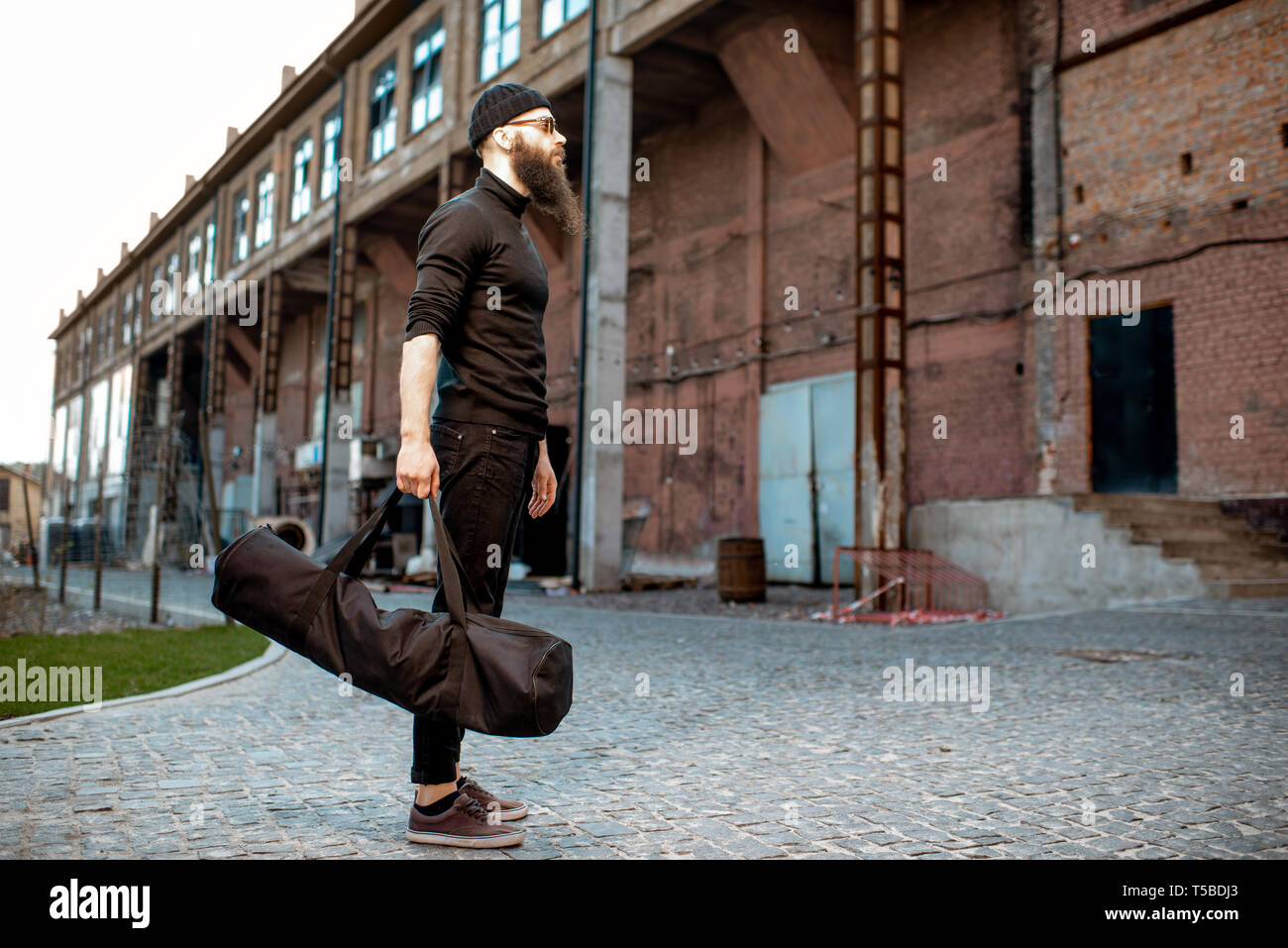 Portrait of a serious bearded man as a killer dressed in black tight clothes holding bag with weapons outdoors on the industrial background Stock Photo