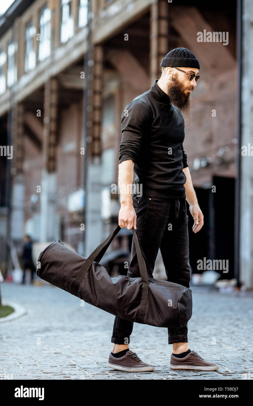 Portrait of a serious bearded man as a killer dressed in black tight clothes holding bag with weapons outdoors on the industrial background Stock Photo