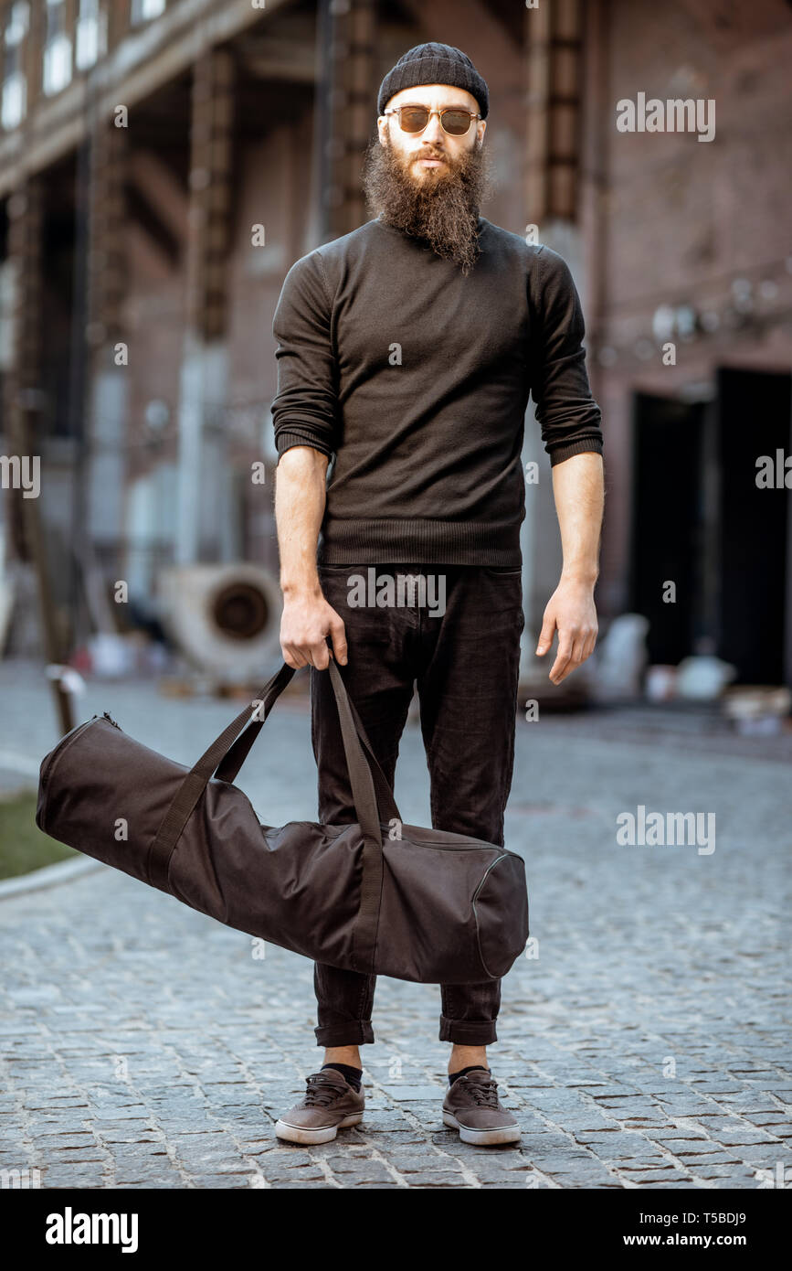 Portrait of a serious bearded man as a killer dressed in black tight clothes holding bag with weapons outdoors on the industrial background Stock Photo