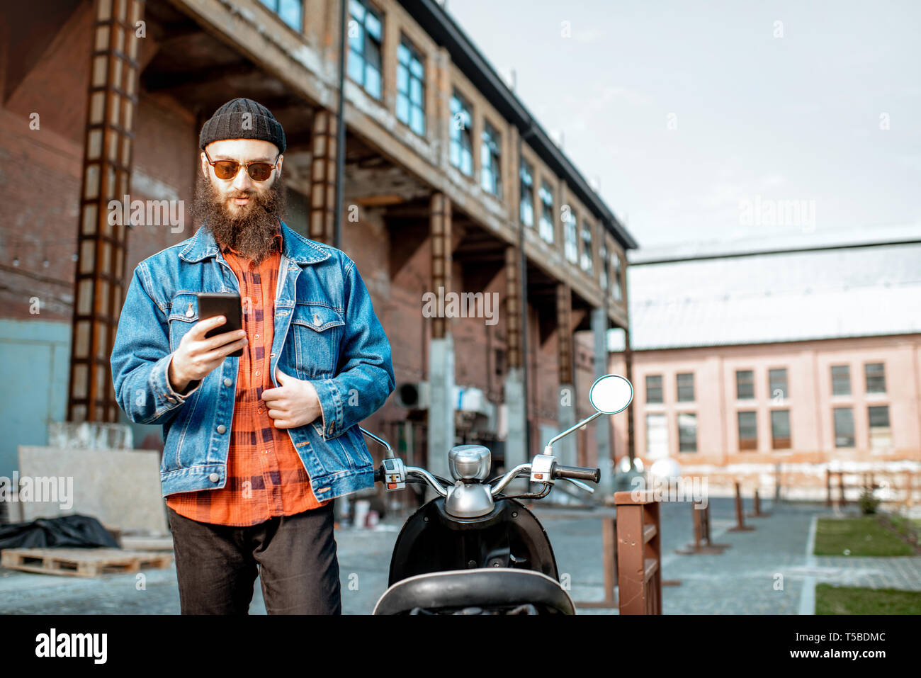Lifestyle portrait of a bearded hipster standing with phone near the retro moped outdoors on the industrial urban background Stock Photo
