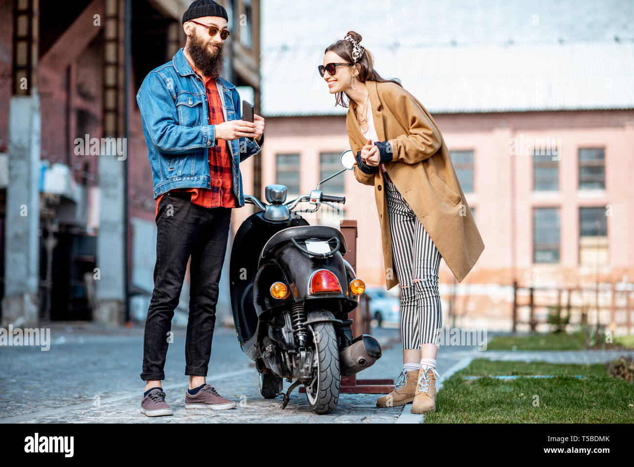 Stylish young man and woman talking together, standing near the retro moped outdoors on the industrial urban background Stock Photo