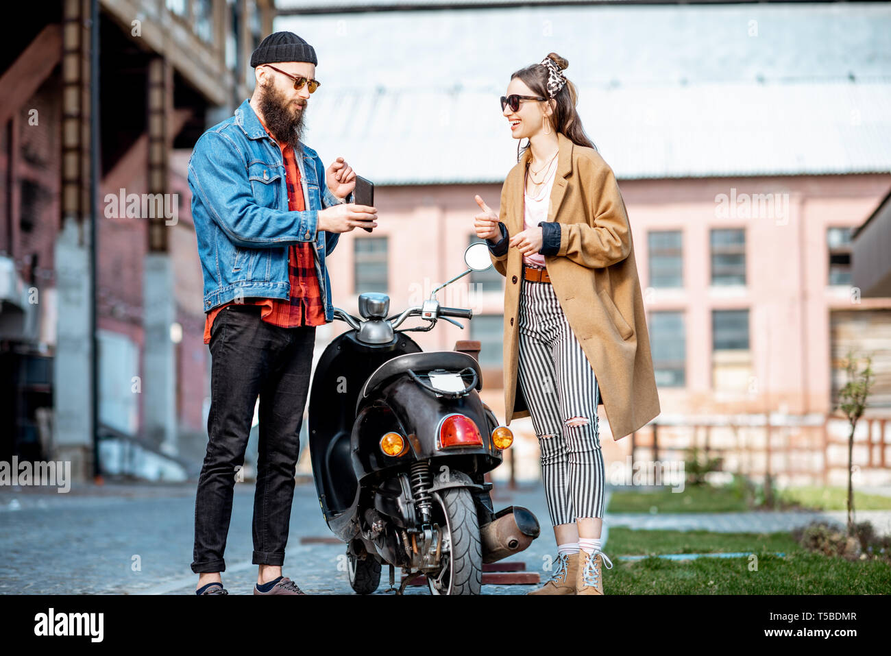 Stylish young man and woman talking together, standing near the retro moped outdoors on the industrial urban background Stock Photo