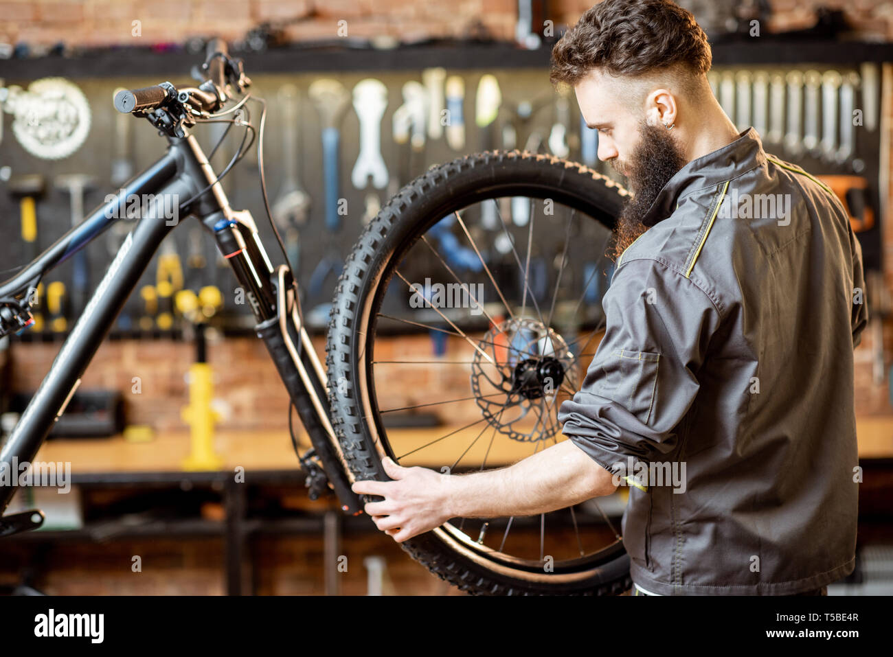 Handsome repairman in workwear serving mountain bicycle, standing with front wheel at the workshop of a bicycle shop Stock Photo