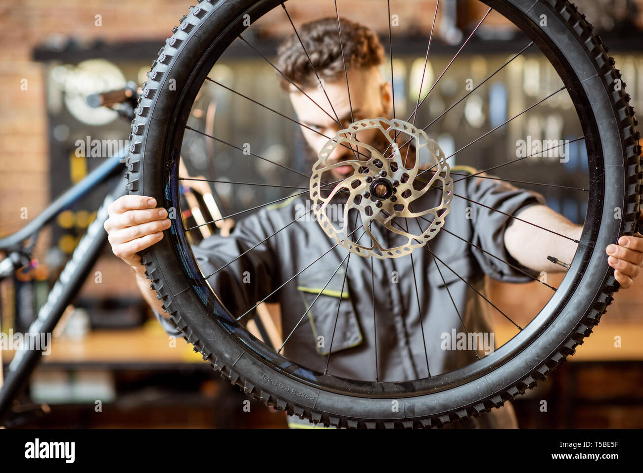Handsome repairman in workwear serving mountain bicycle, standing with front wheel at the workshop of a bicycle shop Stock Photo