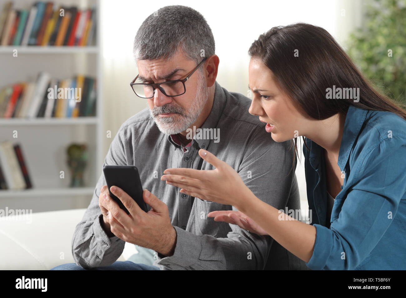 Confused father and daughter trying to use a smart phone sitting on a couch at home Stock Photo