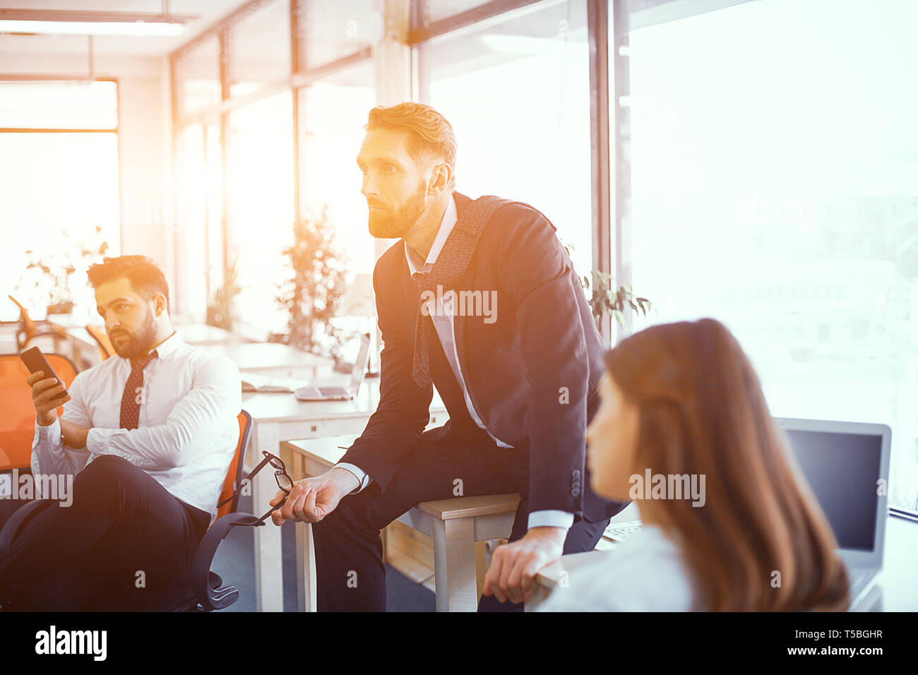 Redhaired Boss Sits On Desk In Office. Coleagues Sitting Nearby