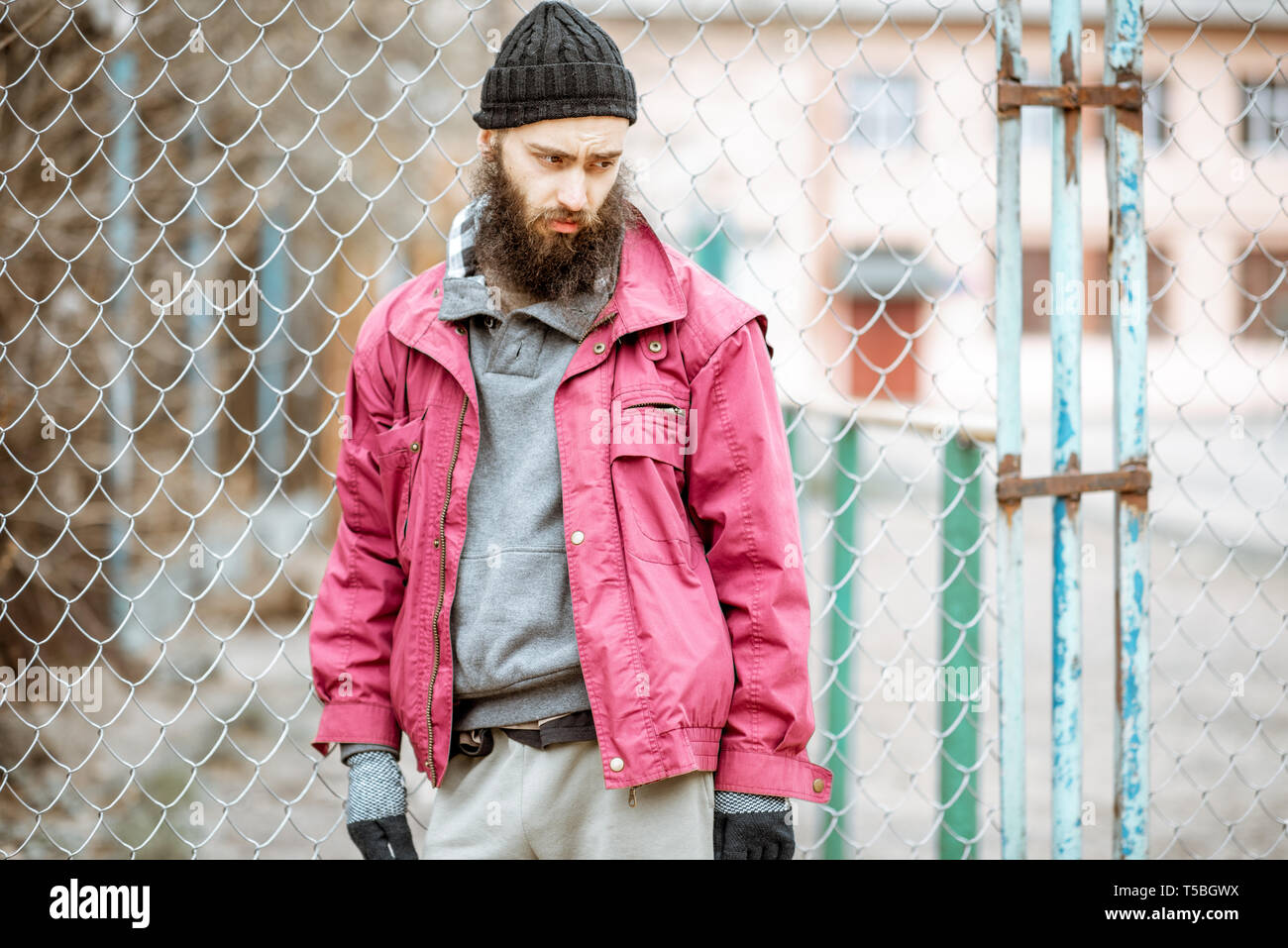 Portrait of a depressed homeless beggar or prisoner standing near the old metal fence outdoors Stock Photo
