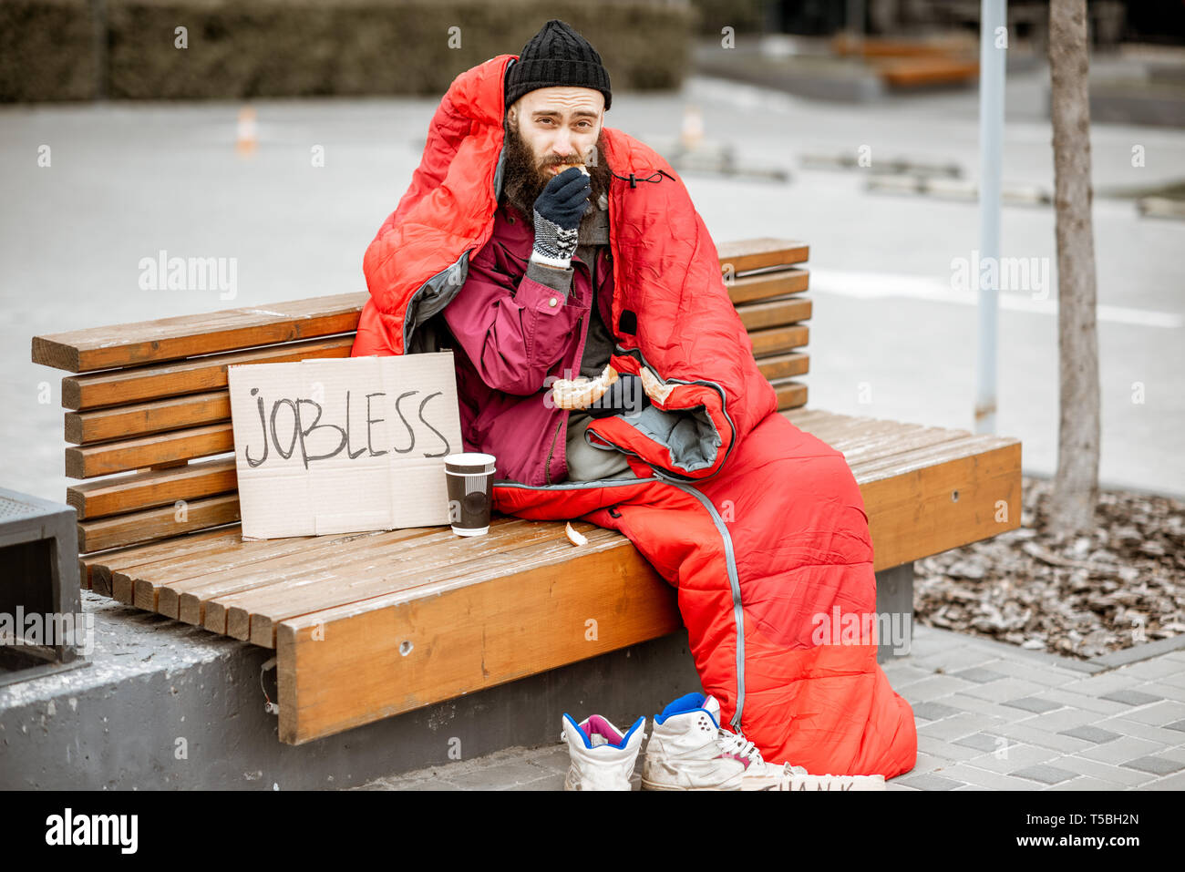 Depressed homeless beggar eating bread while sitting wrapped with