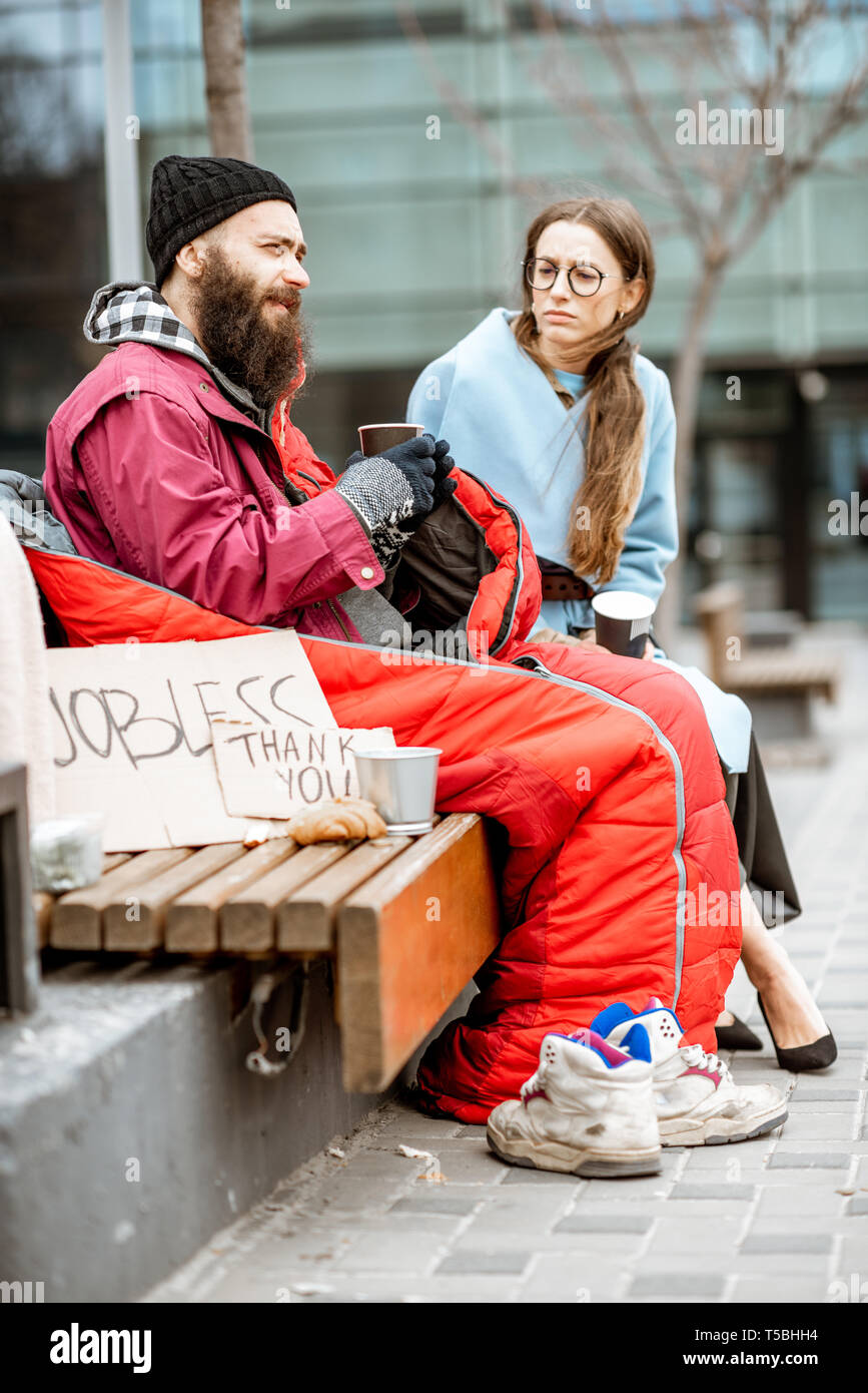 Homeless beggar with young woman listening to his sad story while sitting together on the bench outdoors. Concept of a human understanding Stock Photo