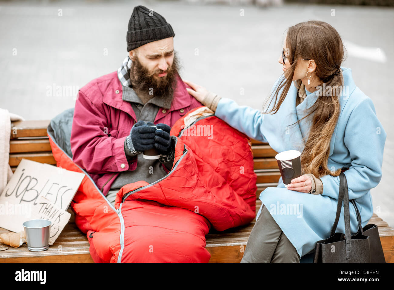Homeless beggar with young woman listening to his sad story while sitting together on the bench outdoors. Concept of human understanding Stock Photo