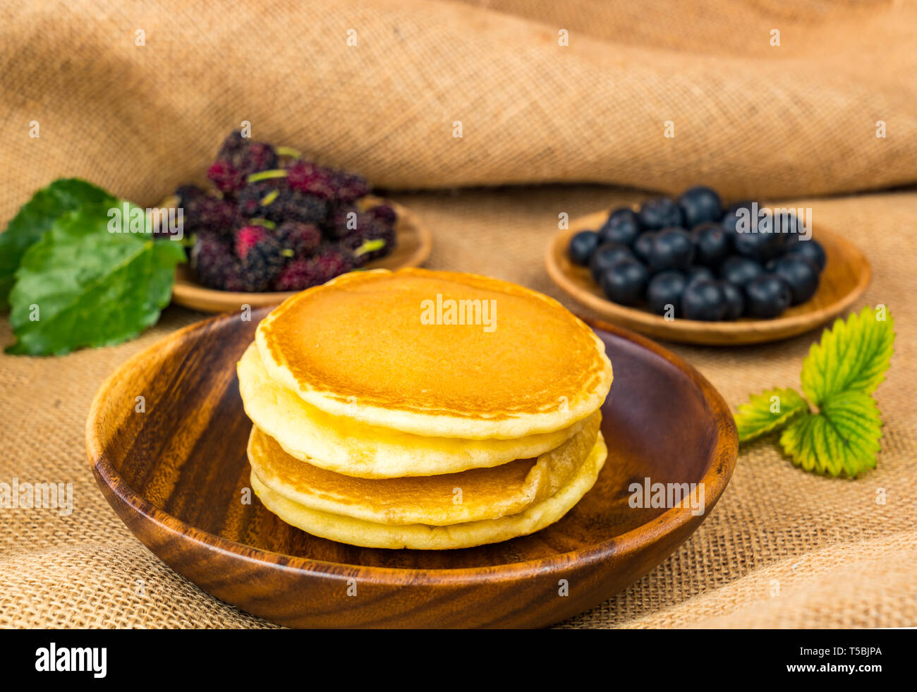 Pile of pancake in wooden bowl with mulberry and blueberry in wooden plate on sackcloth Stock Photo