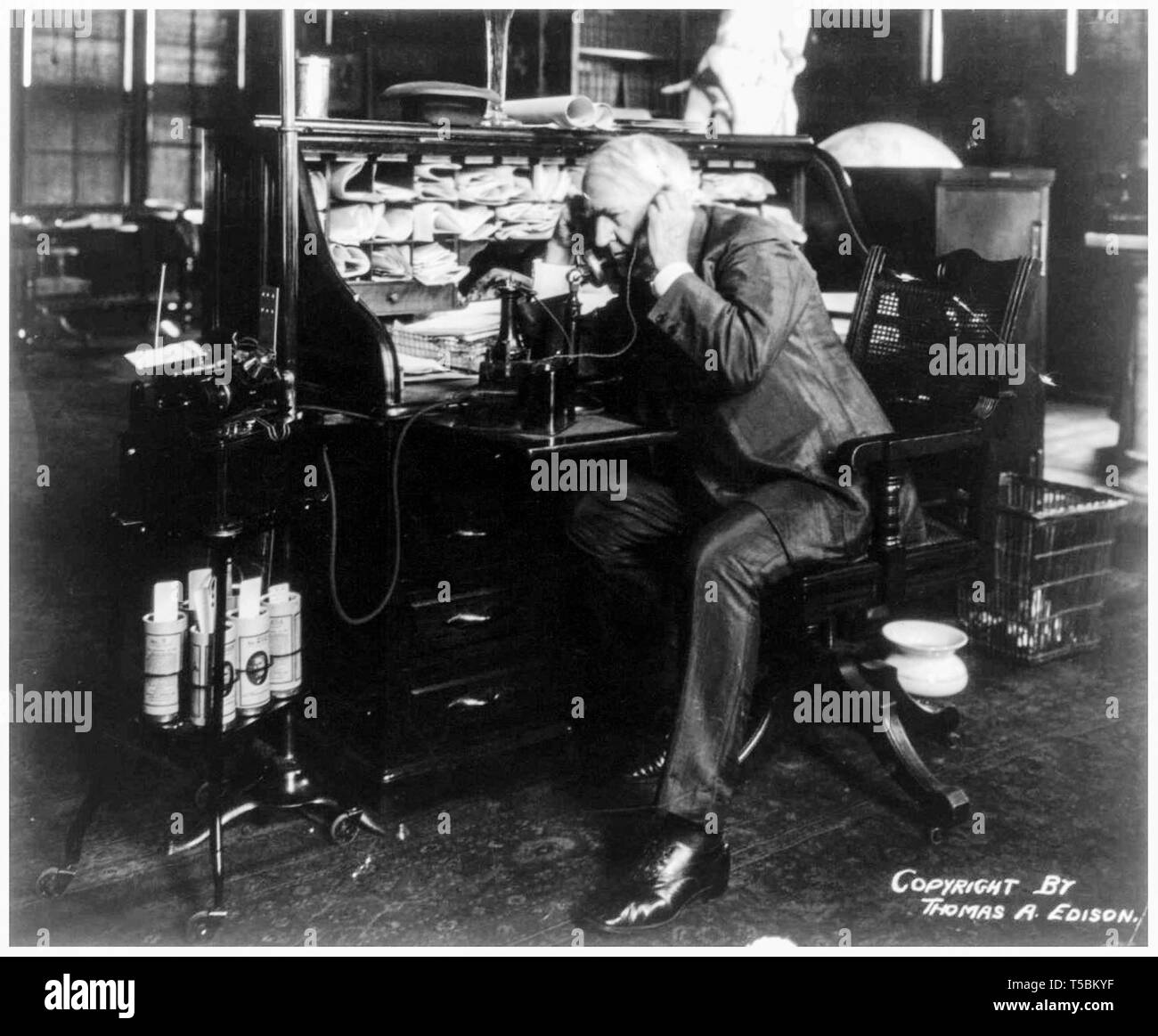 Thomas Edison (1847-1931) at his desk, talking on telephone, showing a wax cylinder dictating machine and cylinders, 1914 Stock Photo