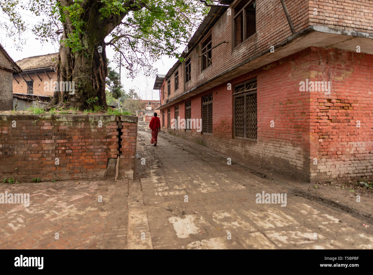 BHAKTAPUR, NEPAL - APRIL 5, 2019: Woman wearing red clothes walking nearby an industrial looking building, taken in an overcast morning in the histori Stock Photo