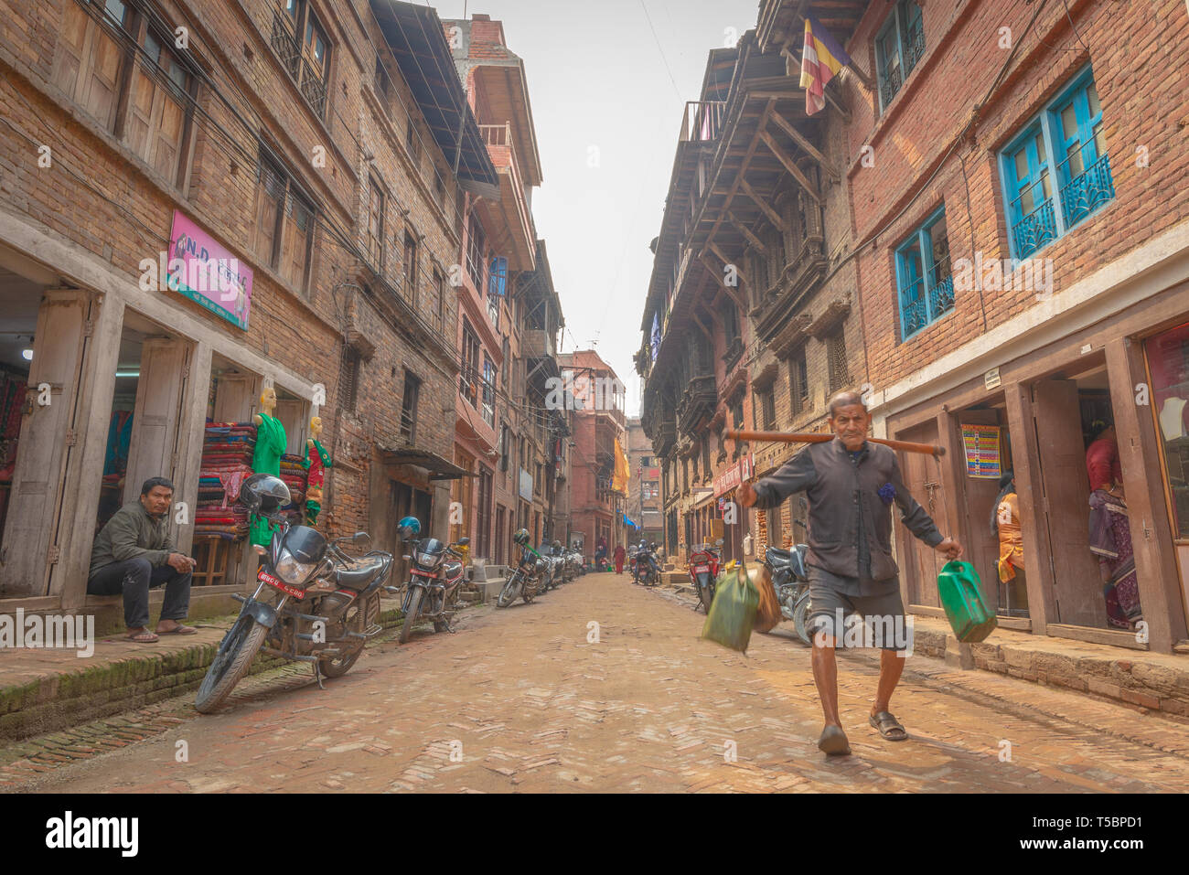 BHAKTAPUR, NEPAL - APRIL 5, 2019: Old man porting plastic cans walking down a narrow street taken during a partly sunny afternoon Stock Photo