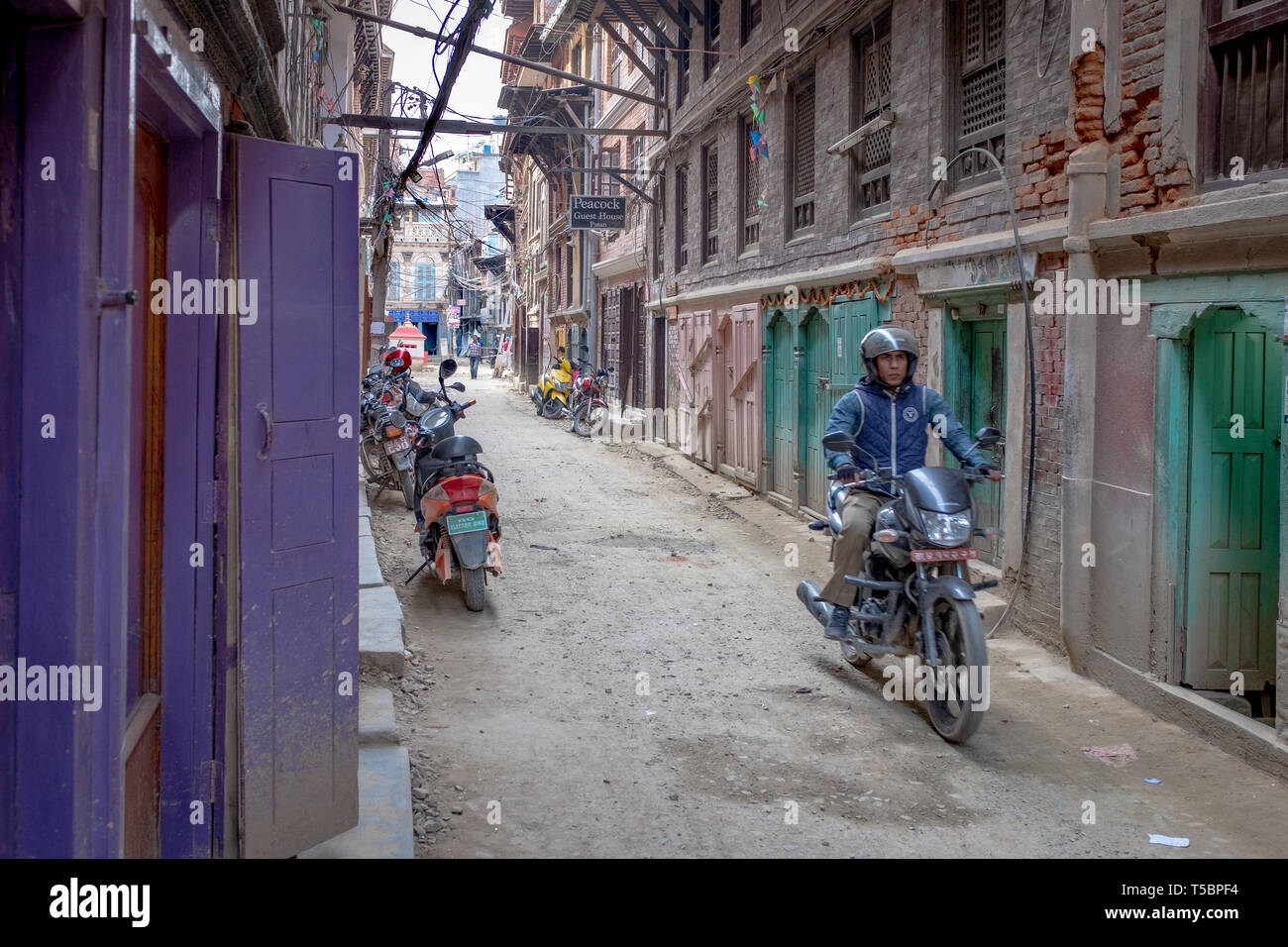 LALITPUR, PATAN, NEPAL - APRIL 3, 2019: A motorcyclist is going down a narrow street lined with colorful doors, taken during a cloudy morning  in the  Stock Photo