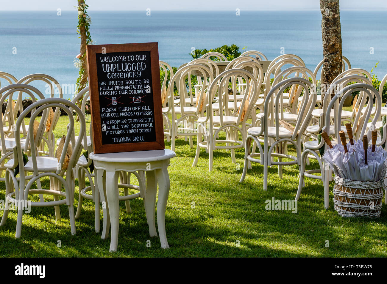 Wedding arch for a ceremony, chairs and sun umbrellas for guests among palm trees near the ocean. Blackboard sign saying to turn cell phones off. Stock Photo