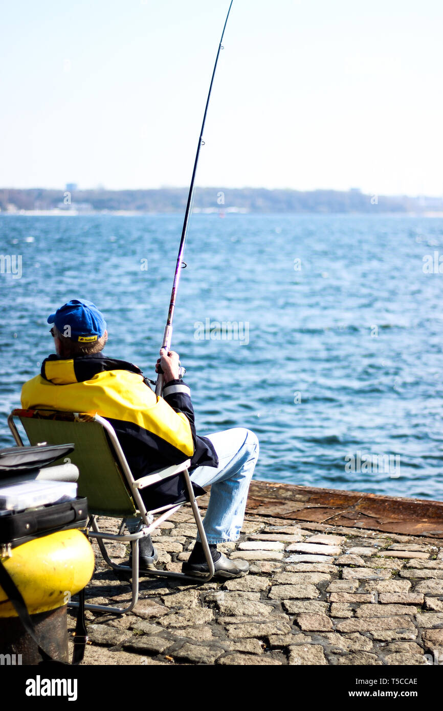 Fischersmann in schwarz-gelber Jacke sitzt an Kieler Förde mit Angel und fängt Fische Stock Photo