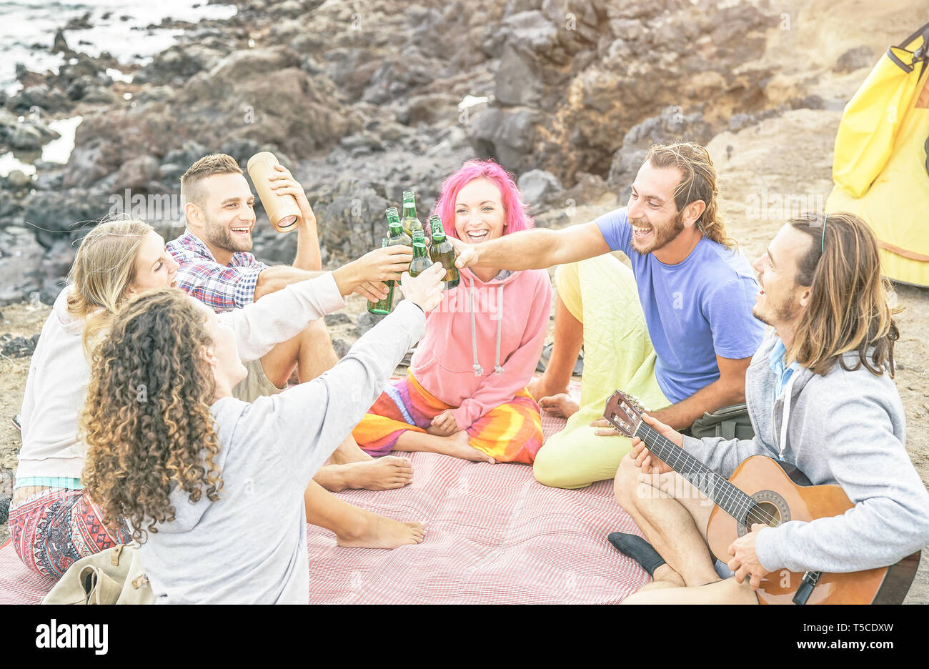 Group of happy friends toasting beers and playing guitar camping with tent on the beach - Travel people having fun listening music and drinking Stock Photo