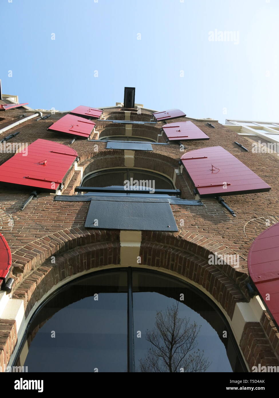Looking upwards at the distinctive style of a tall canal-side building in Amsterdam with arched windows and red shutters folded to the sides. Stock Photo