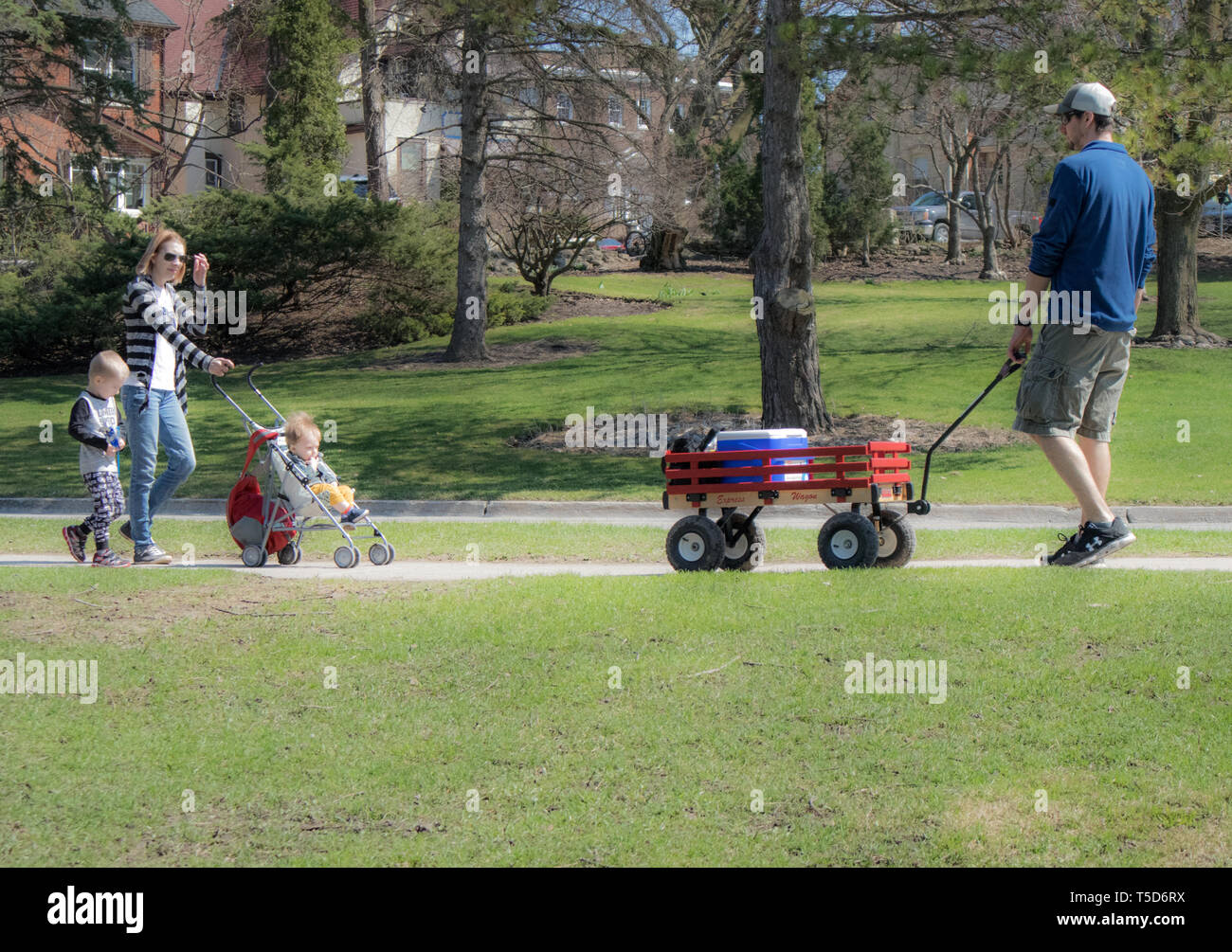 Young couple with very young kids, walking in the park during a beautiful spring day. Dad pulls a trolley, while mum pushes a stroller. Stock Photo
