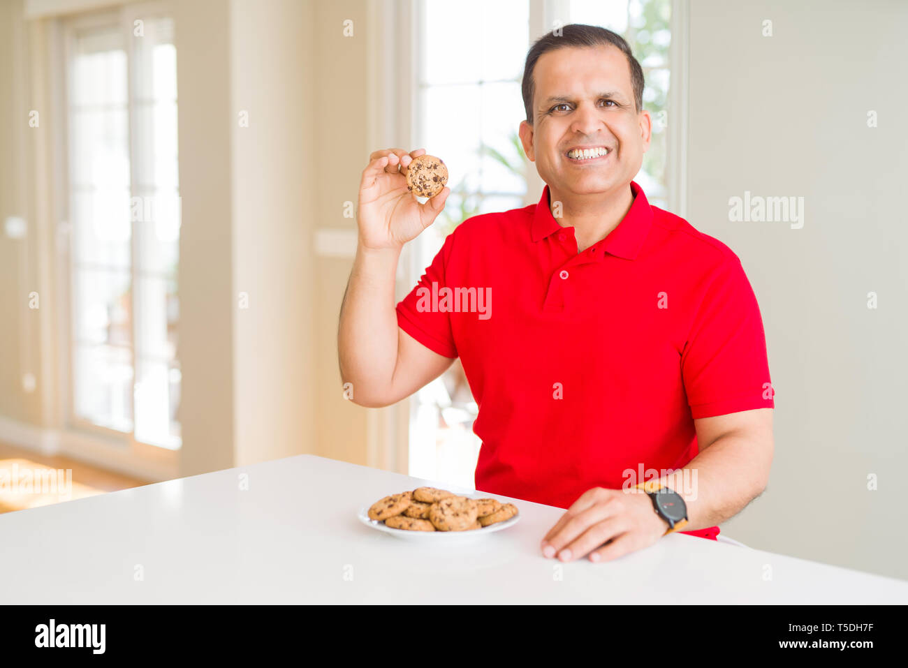 Middle age man eating chocolate chips cookies at home with a happy face standing and smiling with a confident smile showing teeth Stock Photo