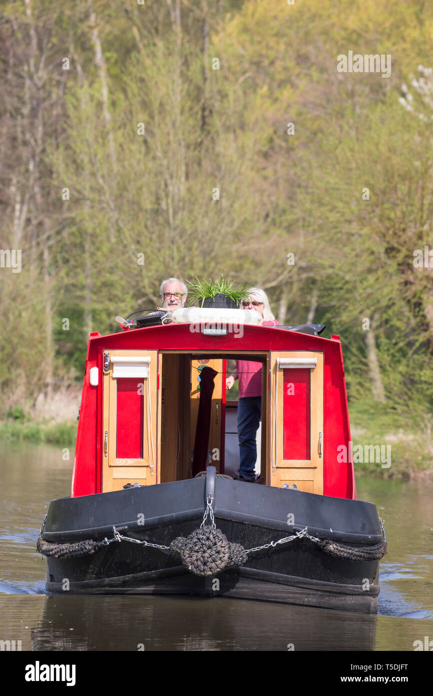 Portrait shot of oncoming narrowboat on UK canal, a retired married couple at rear of the boat, facing forwards, steering bright red canal boat in sun. Stock Photo