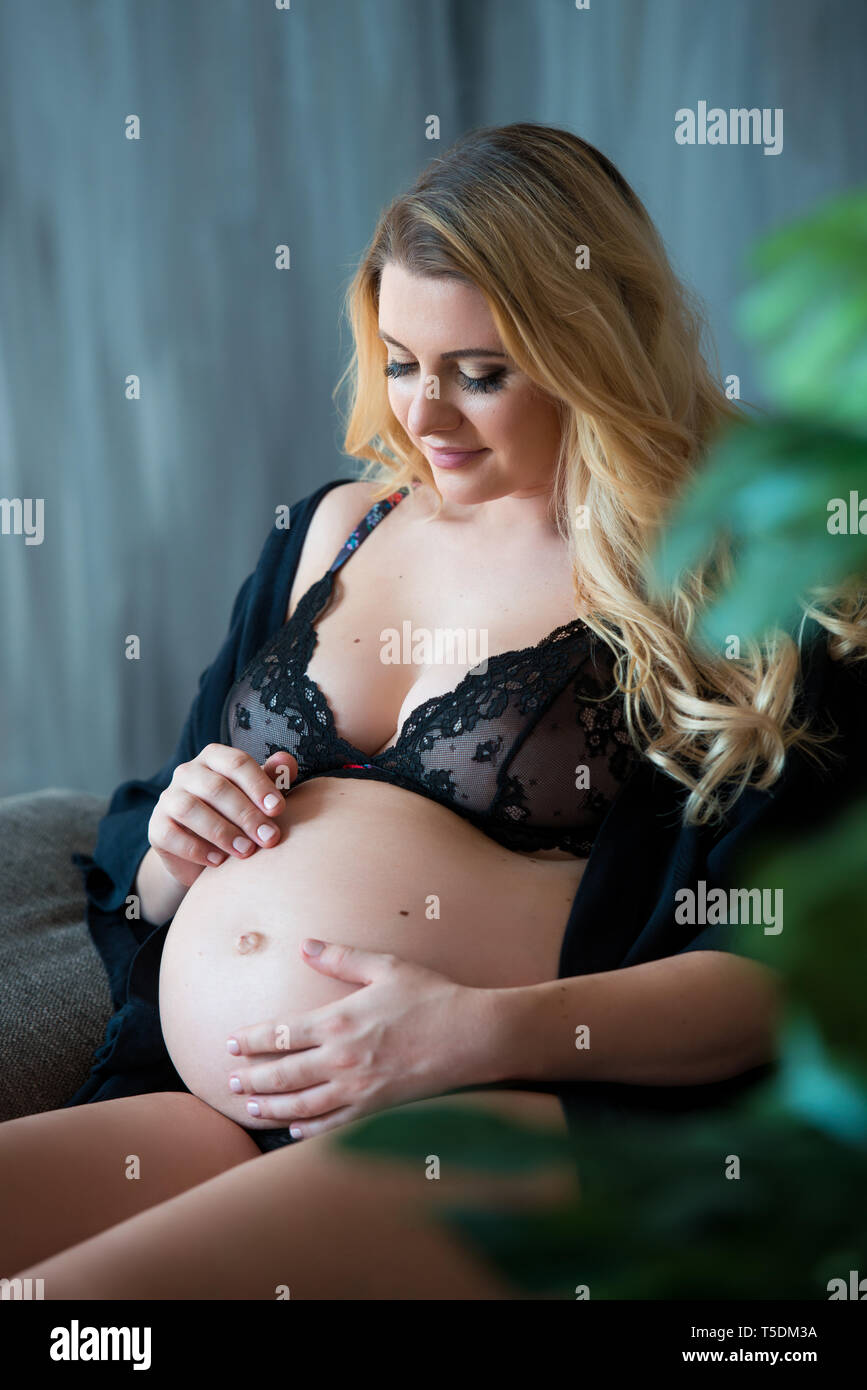 Pregnant photoshoot in loft style. Beautiful pregnant mother with blond hair  sits on sofa and dreamily looks out the window and on tummy Stock Photo -  Alamy